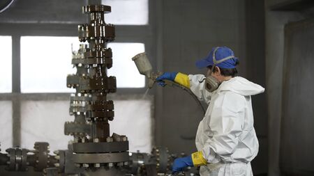 Worker in protective respirator and white suit paints oil and gas wellhead tree equipment with spray gun in spray booth. Equipment for wellhead conncetion of oil and gas wells.の写真素材