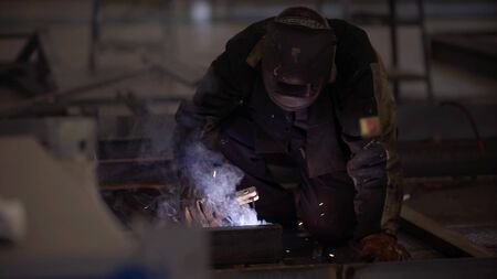 Blacksmith welder in protective mask works with metal steel and iron using welding machine, bright...
