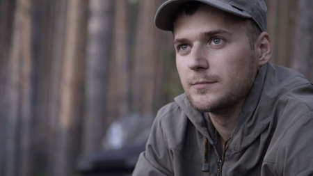 Portrait of young adult male tourist sits on log near fire in forest. Human looks at forest fire on nature. Cooking dinner on wild hike. Outdoor travel. Close up shotの写真素材