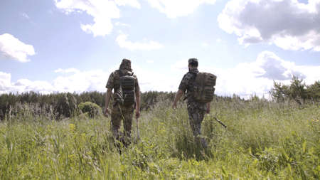 Rear view of two men walking with fishing poles in field to river. Relaxed men spending leisure...