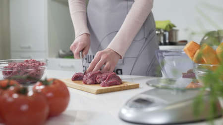 Woman cutting raw meat on small pieces with kitchen knife cutting board. Woman puts pieces of raw meat in a glass bowl. Female cutting red beef filet on wooden board, holding knife in hand.の写真素材