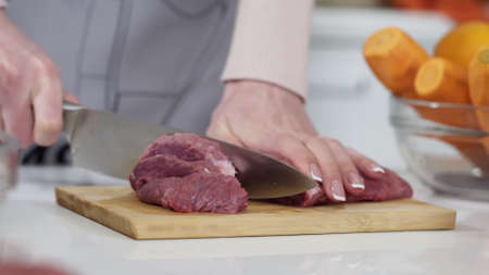 Woman cutting raw meat on small pieces with kitchen knife cutting board. Female cutting red beef filet on wooden board, holding knife in hand. Housewife is cooking at home interior. Concept: foodの写真素材