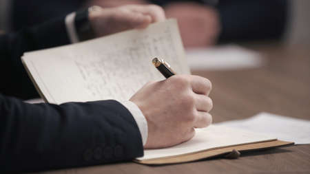 Male hand holding pen and writing to do list or business data with pen at notebook dairy. Hand writing business report, make note on paperwork document on office desk in corporate meeting room.の写真素材