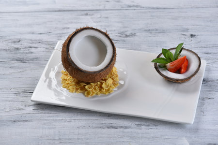 coconut dessert in coconut with pulp, corn flakes and strawberries on a white square plate, pina...