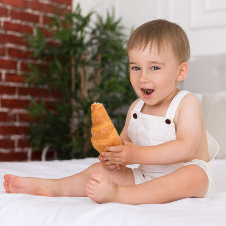 happy portrait of a little smiling boy child playing and eating a croissant sitting on the bedの写真素材