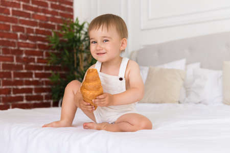 happy portrait of a little smiling boy child playing and eating a croissant sitting on the bedの写真素材