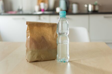Brown paper lunch bag and bottle of water on wooden table over kitchen background. Paper bag with food. Lunch time.の写真素材