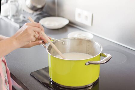 Woman preparing bechamel sauce or cream in a pan. Woman hand is mixing boiling milk with wooden spoon.の写真素材