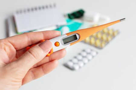 Female hand holding a digital thermometer on white background with pills. Cold and flu concept.の写真素材