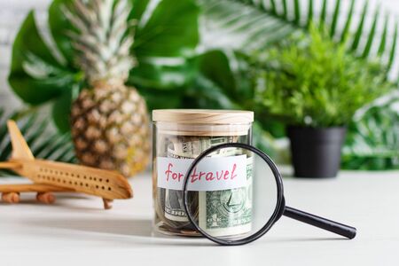 Money savings for travel in a glass jar on white wood table over wall with green plant leaves . Savings for travel budget concept.の写真素材