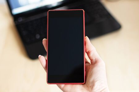 Woman hand holding a smartphone with black blank screen against table with laptop. Business, technology concept. Mockup for design.の写真素材