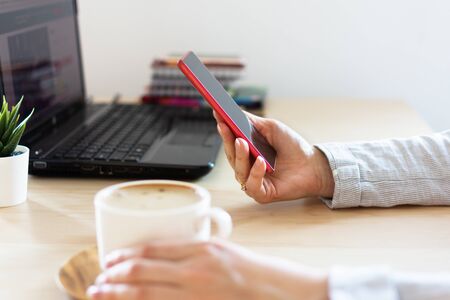 Woman hand holding a smartphone against table with laptop in office. Coffee break. Business concept. Freelance or blogging.の写真素材