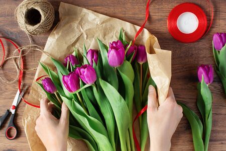 Woman florist wrapping bouquet of amazing purple tulips in craft paper on wooden table top view.の写真素材