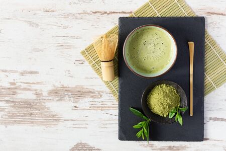 Green matcha tea drink and tea accessories on black board on white wooden table top view. Japanese tea ceremony concept. Chashaku spoon and chasen bamboo whisk for brewing matcha tea.の写真素材