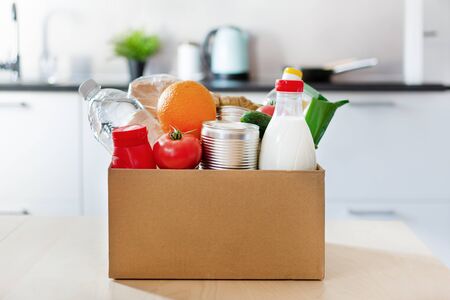 Cardboard box with various food, bottle of milk and water on the table over kitchen interior. Food donations or food safe delivery concept. Safe home delivery.の写真素材