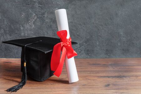 Academic cap and graduation diploma scroll and tied with red ribbon on wooden table over black chalkboard.の写真素材