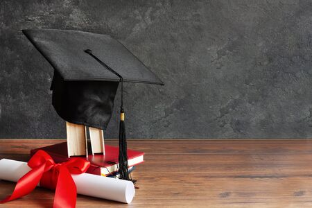 Academic cap and graduation diploma scroll and tied with red ribbon on wooden table over black chalkboard.の写真素材