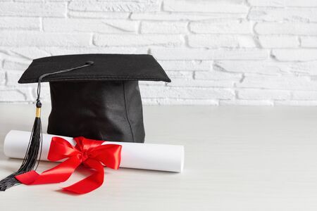 Academic cap and graduation diploma scroll and tied with red ribbon on white wooden table.の写真素材