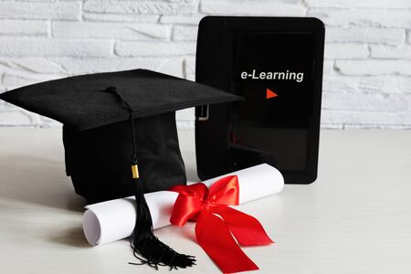 Academic cap and graduation diploma scroll and tied with red ribbon and tablet on white wooden table. E-learning concept. Remote studying.の写真素材