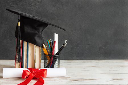 Academic cap and graduation diploma scroll and tied with red ribbon on white wooden table over black chalkboard.の写真素材