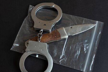 Knife in a plastic package and handcuffs on black color background table. Physical evidence pack. Criminal concept.の写真素材