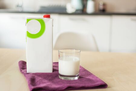 A glass of milk with napkin and paper bottle on wooden table over blur white kitchen background. Blank paper container with milk for design. Organic milk concept.の写真素材