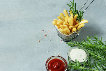 French fries in basket with ketchup, sauce and rosemary on grey background. Copy space.