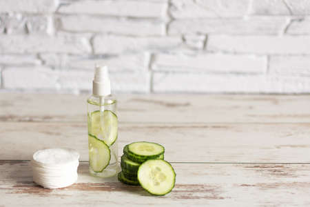 Homemade refreshing facial cucumber toner in bottle with slices of green cucumber and cotton-pads on white wooden background. Copy space.の写真素材