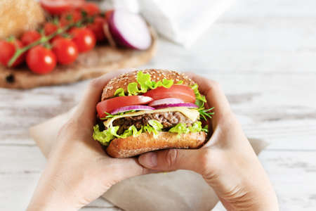 Woman hands hold beef burger with fresh salad, cheese and tomatoes over table. Fast food, unhealthy eating.の写真素材