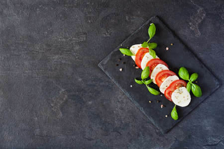 Caprese salad with mozzarela, tomatoes, fresh olive oil and basil on slate board on black background...