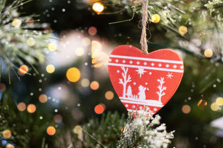 Christmas toy in the shape of a red heart hanging on the Christmas tree with beautiful bright bokeh of garland. Christmas greeting card with copy space. Christmas or New Year background.の写真素材