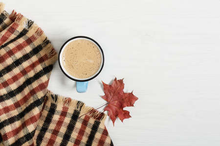 Cup of coffee, plaid and dried maple leaf on white wooden background. Autumn composition.