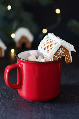 Red mug of hot chocolate with marshmallow and homemade gingerbread small house for decoration on...