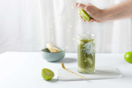 Woman hand squeezing lime into glass with ice green matcha latte on white background. Iced matcha lemonade.の写真素材
