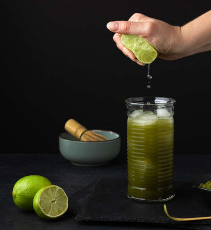 Woman hand squeezing lime into glass with ice green matcha tea on black background. Still life of iced matcha lemonade.の写真素材