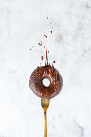 Donut on fork with splashing chocolate glaze over grey background. Sweet food advertising, creative picture of sweet chocolate dessert.の写真素材