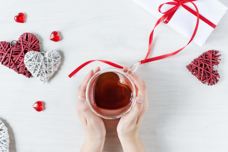 Woman hands holding tea cup in a heart shaped with red hearts decoration on white wooden table top view. St. Valentine's Day concept. Valentine's Day romantic dinner.の写真素材