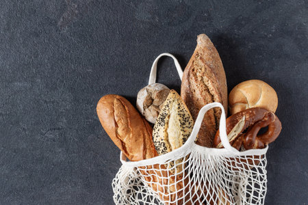 Assortment of baked bread and buns in white mesh tote bag on black  background top view. Healthy organic bread. Eco-shopping concept.の写真素材