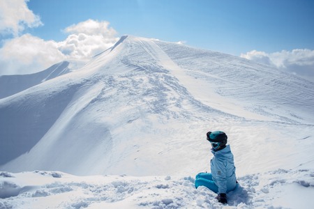 Young girl skier sits on the edge of the hillside and looking at the snow-covered mountain peak in sunny clear winter dayの写真素材