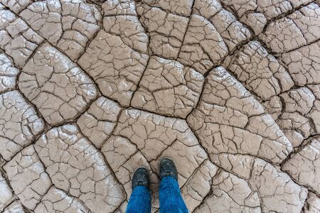 Feet on a dry and cracked ground surface. Boots and salt polluted soil. Climate change. Environmental pollution by chemical waste. Ecological catastrophyの写真素材