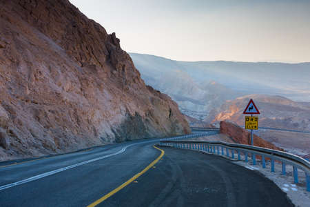 Road in the Negev Desert in Israel. Two-lane Desert Highway. Road signs Slow in English, Arabic and Hebrew and sharp turn. Trail among rocks and blue sky.の写真素材