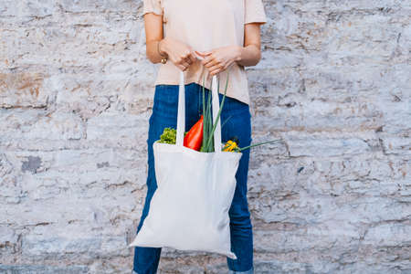 Woman holding white canvas tote bag with vegetables near brick wall. Reusable eco concept.の写真素材