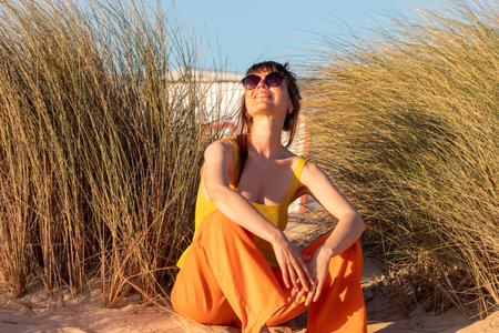 A beautiful woman in orange clothes sits on the sand surrounding by beach grass.の写真素材