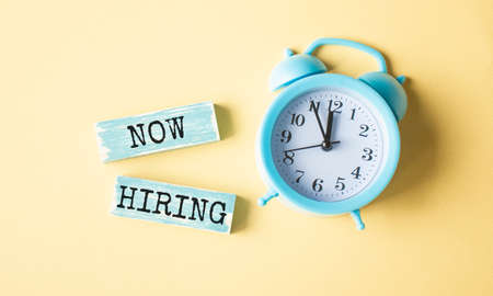 New Hiring text written on wood block. Now we are hiring text on the yellow table with a blue alarm clock, for your design.の写真素材