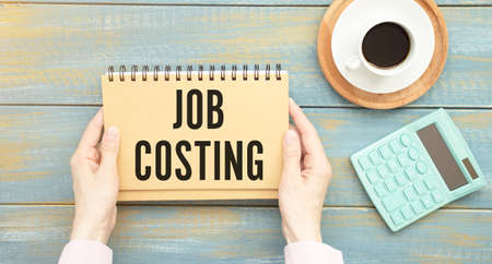JOB COSTING text on paper sheet with chart, dice, spectacles, pen, laptop and blue and yellow push pin on wooden table - business, banking, finance and investment concept. BUSINESS CONCEPTの写真素材