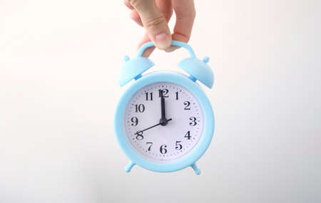 hand holding white alarm clock. Finger pointing to arrow of twelve o'clock. Light pastel blue background. Time change concept. Closeup.の写真素材