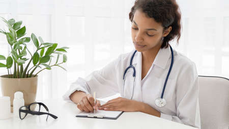 African american smiling female doctor sitting at the table in the medical office and working on the...