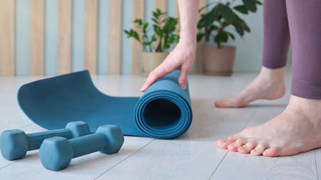 A barefoot woman twists a purple yoga Mat and fitness on the parquet floor.の写真素材