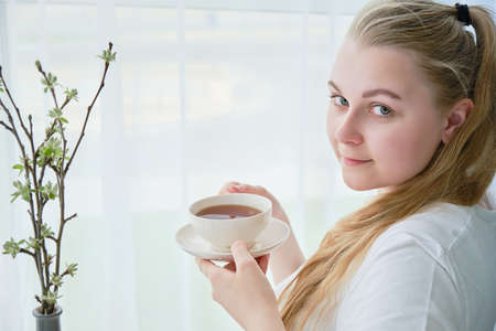 Young woman with a mug of tea in her hands sits near the window, space for text.