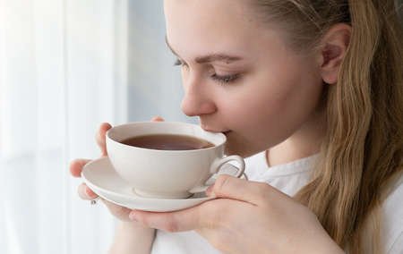 Young woman with a mug of tea in her hands sits near the window, space for text.の写真素材